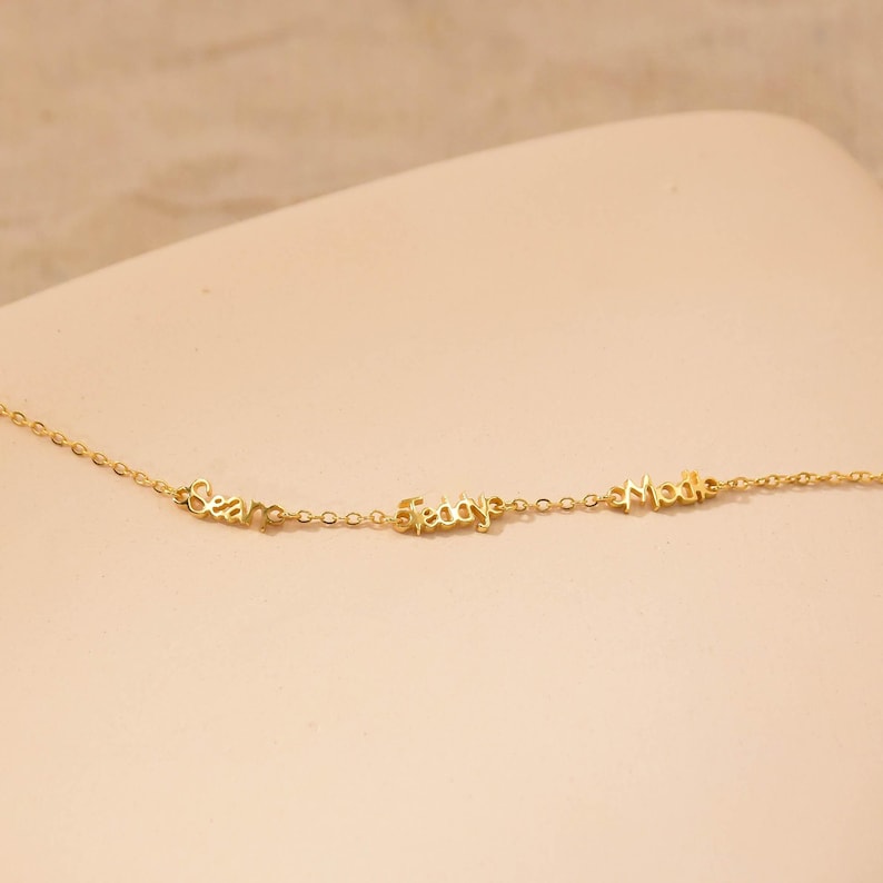 May include: A delicate gold necklace with three cursive name pendants: "Sean", "Fady", and "Madi". The chain and pendants are a warm gold color, creating a simple yet elegant design. The necklace is displayed on a neutral background.