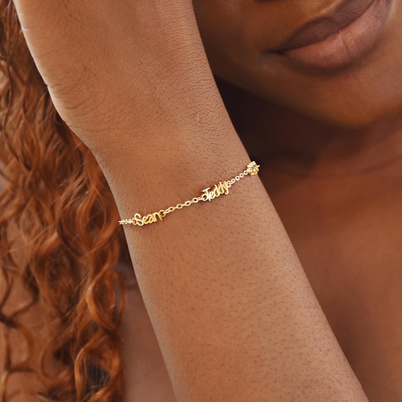 May include: A delicate gold bracelet with the names "Sean" and "Teddy" spelled out in cursive script. The bracelet is connected by a fine chain and is worn on a wrist. The bracelet is a personalized jewelry item.