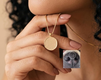 Könnte beinhalten: Eine goldfarbene Halskette mit einem runden Anhänger, dessen strukturierte Oberfläche das Muster einer Hundenase nachahmt. Der Anhänger hängt an einer feinen Boxkette. Ein eingefügtes Bild zeigt eine Nahaufnahme einer schwarzen Hundenase.