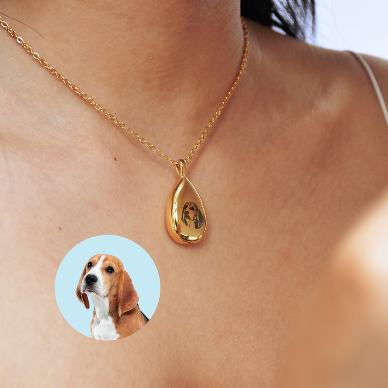 May include: A gold chain necklace with a teardrop-shaped pendant featuring a black and white photo of a dog. The pendant is about 1 inch long.