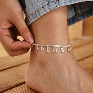 May include: Silver anklet with dangling letters spelling "I PLAS" and a heart charm. The anklet is worn on a foot, with a hand adjusting the clasp. The letters and heart are embellished with small, sparkling stones.