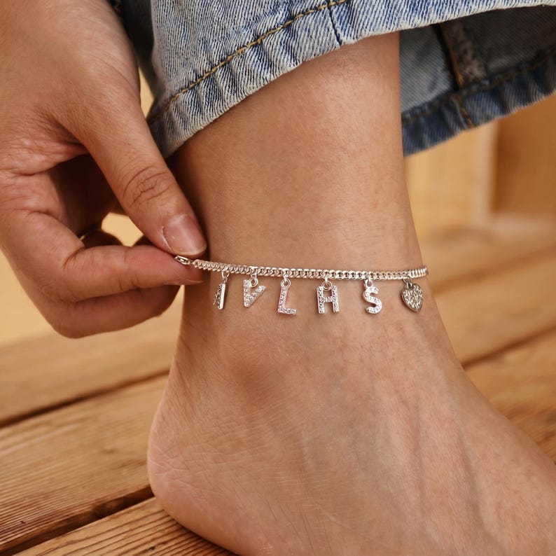 May include: Silver anklet with dangling letters spelling "I PLAS" and a heart charm. The anklet is worn on a foot, with a hand adjusting the clasp. The letters and heart are embellished with small, sparkling stones.