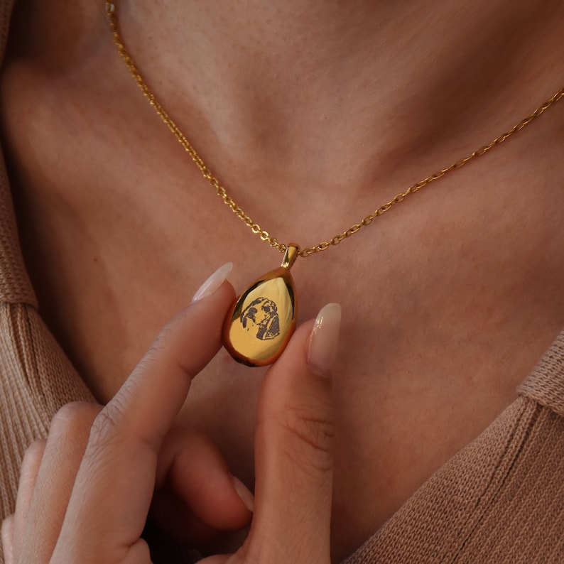 May include: A gold teardrop-shaped pendant necklace with a black and white photo of a dog engraved on the front.