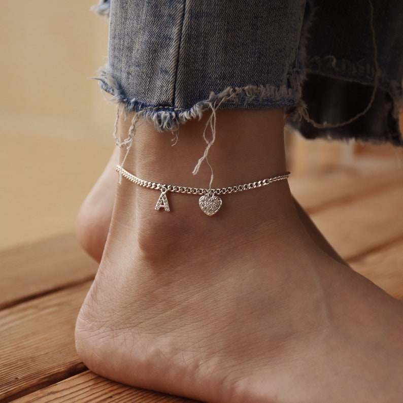 May include: Silver anklet with a curb chain design. The anklet features the letter "A" charm and a heart-shaped charm. The anklet is worn on an ankle with a pair of blue jeans in the background.