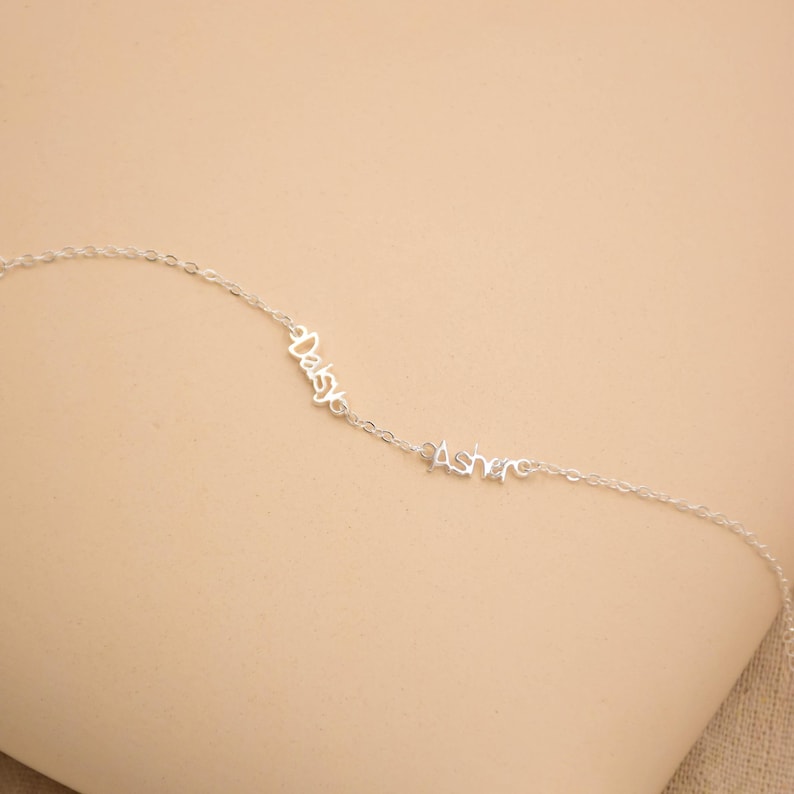 May include: A delicate silver necklace with the names "Daisy" and "Asher" in cursive script. The necklace features a fine chain and is displayed against a neutral background. A personalized jewelry item.