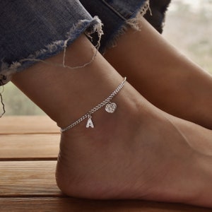 May include: Silver anklet with a chain design, featuring a charm with the letter "A" and a heart-shaped charm. The anklet is worn on an ankle, with denim jeans visible in the background.