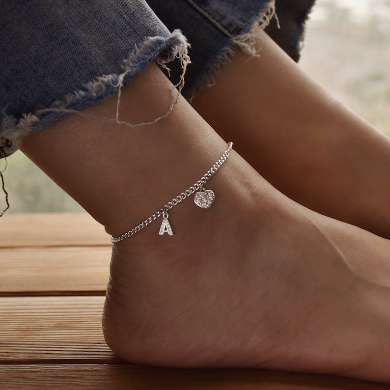 May include: Silver anklet with a chain design, featuring a charm with the letter "A" and a heart-shaped charm. The anklet is worn on an ankle, with denim jeans visible in the background.