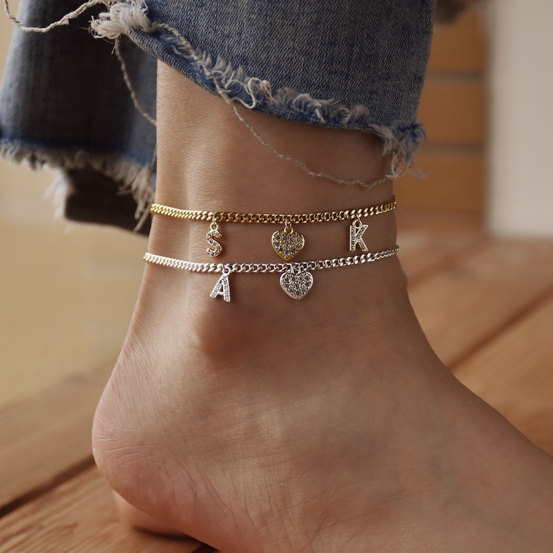 May include: Two anklets with chain links and dangling charms. One gold-toned anklet has a letter "S" charm and a heart charm. The other silver-toned anklet has a letter "A" charm and a heart charm. The anklets are worn on an ankle.