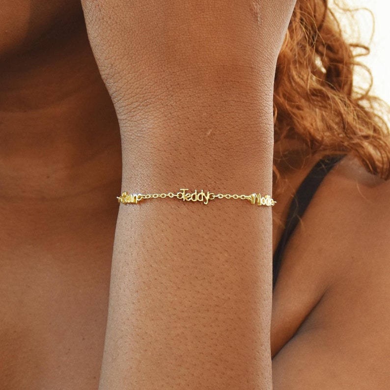 May include: A delicate gold-colored bracelet with the names "Liam", "Teddy", and "Mollie" spelled out in cursive script. The bracelet is worn on a person's wrist, showcasing the personalized jewelry.