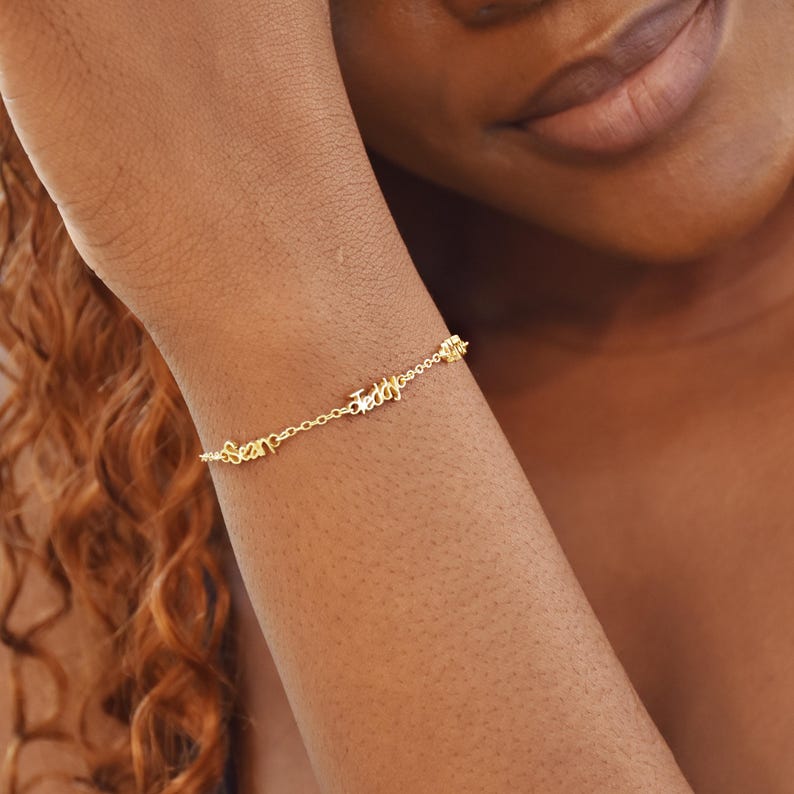 May include: A delicate gold bracelet with the names "Sean" and "Teddy" spelled out in cursive script. The bracelet is attached to a chain and is worn on a wrist. The bracelet is a piece of jewelry.
