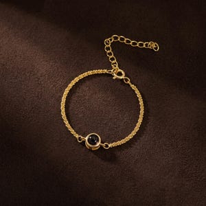 以下が含まれることがあります： テクスチャードチェーンと中央に小さな丸い黒い石が付いたゴールドカラーのブレスレット。ブレスレットには、サイズ調整可能なチェーンとクラスプクロージャーが付いています。ジュエリーは濃い茶色の表面に表示されています。