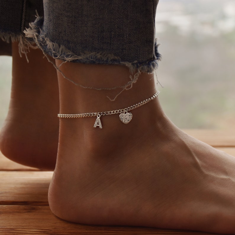 May include: A silver anklet with a chain design. The anklet features a small letter "A" charm and a heart-shaped charm. The anklet is worn on an ankle, with a blurred background.