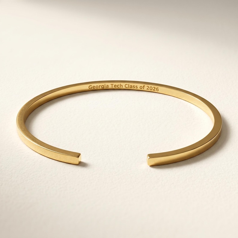 May include: A gold-colored open cuff bracelet. The bracelet has the engraved text "Georgia Tech Class of 2026" on the inside. The bracelet is made of a shiny, smooth metal.