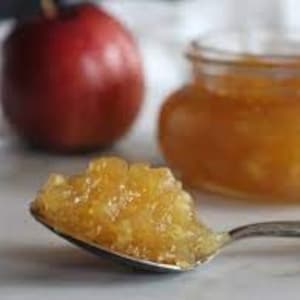 May include: A close-up of a spoonful of golden apple butter, with a whole apple and a jar of apple butter in the background.
