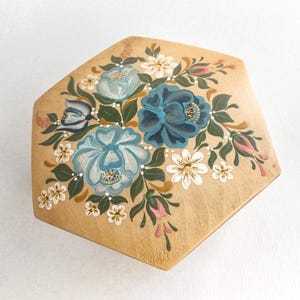 May include: Hexagonal wooden box featuring a hand-painted floral design. The flowers are in shades of blue, white, and pink, set against a light brown background. Green leaves and white dots accent the floral arrangement.