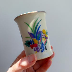 May include: A cream-coloured ceramic vase with a flared top, decorated with a vibrant floral design. The flowers are painted in shades of red, yellow, blue, and purple, with green leaves. The vase is held in a hand.