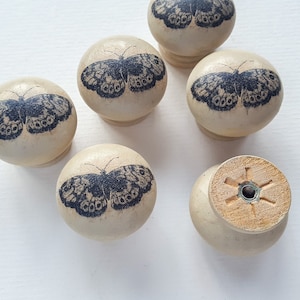 May include: Six beige wooden drawer knobs with black butterfly designs. The knobs have a smooth, rounded surface and a small hole in the centre for attaching to a drawer.
