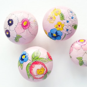 May include: Four pink, spherical decorative knobs with floral designs. Each knob features a different arrangement of colourful flowers, including blue, yellow, pink, and white blooms with green leaves. The knobs are set against a white background.