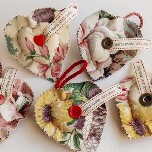 May include: Heart-shaped fabric ornaments with floral patterns in cream, pink, and yellow. Each has a button and a ribbon that reads "hand made with love". Red cord loops are attached for hanging.