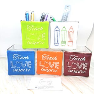 May include: Four glass pencil holders with different coloured backgrounds. The holders are decorated with white outlines of a ruler, apple, pencils, and the words "Teach Love Inspire".
