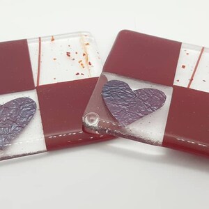 May include: Two square glass coasters with a red and white chequered pattern. Each coaster has a purple heart in the centre.