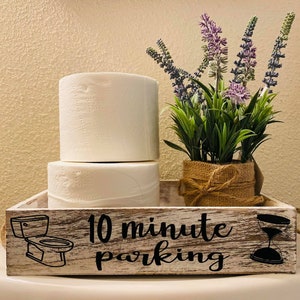 May include: A white wooden tray with the text "10 minute parking" and a black and white illustration of a toilet. The tray has a brown rope handle and is decorated with a burlap wrapped vase with lavender flowers.