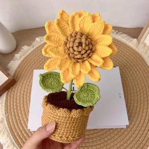 May include: A crocheted sunflower in a small pot. The sunflower has bright yellow petals and a brown centre. The pot is a light brown colour, and the leaves are green. The flower is a decorative item.