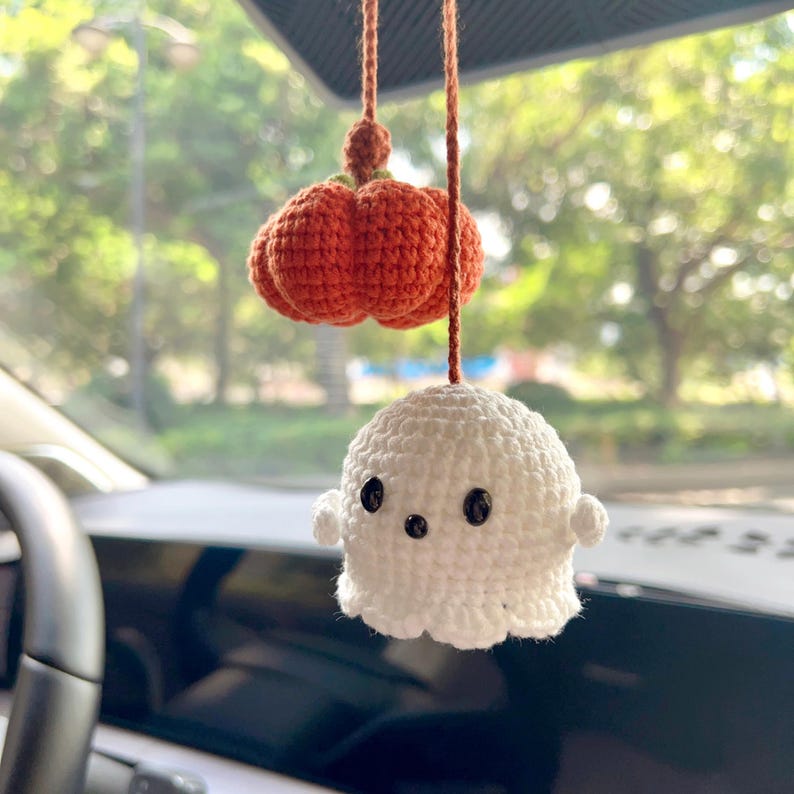 May include: A handmade crochet car charm featuring a small orange pumpkin and a white ghost. The pumpkin is a vibrant orange color, and the ghost has black button eyes. The charm is suspended by a brown string, perfect for Halloween.