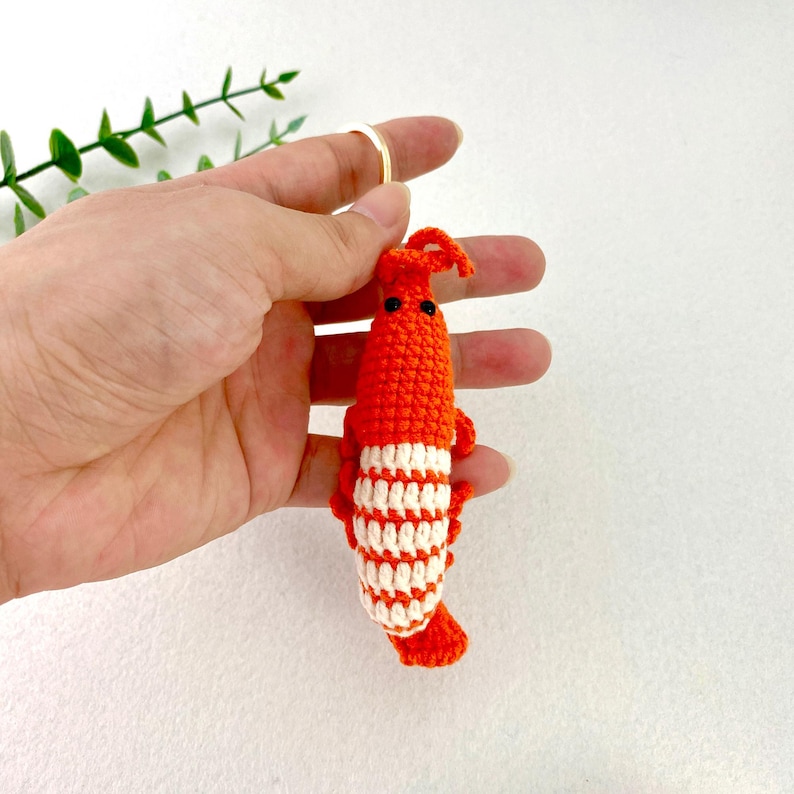 May include: A crocheted shrimp keychain in bright orange and white. The shrimp has a segmented body with white stripes and black eyes. The keychain is held by a gold ring.