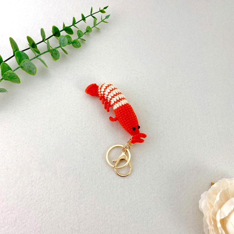 May include: A handmade crochet shrimp keychain in bright red and white stripes. The shrimp has small black eyes and a gold-colored clasp. The keychain is a unique accessory.