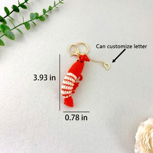 May include: A red and white crocheted shrimp keychain, 3.93 inches long and 0.78 inches wide, with a gold-colored keyring and a small letter "D" charm. The text "Can customize letter" is also visible.