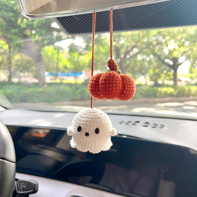 May include: A handmade Halloween car charm featuring a white ghost and an orange pumpkin, both crocheted. The ghost has black button eyes and small arms. The pumpkin is made of three connected sections. The charm hangs from a brown string.