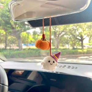 May include: Crocheted Halloween car charm featuring a small orange pumpkin and a white ghost wearing a purple and yellow witch hat. The charm hangs from a brown string, perfect for seasonal decor.