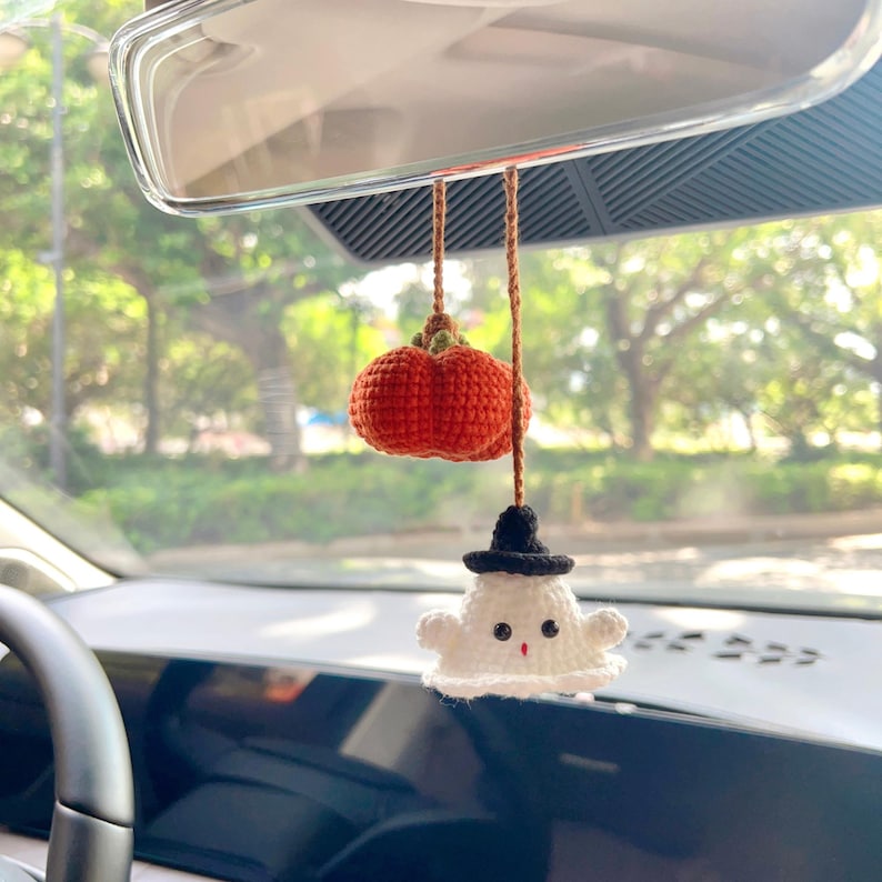 May include: A handmade Halloween car charm featuring a crocheted orange pumpkin and a white ghost with a black hat. The charm hangs from a brown string, adding a festive touch to the car's rearview mirror. The charm is a cute, seasonal decoration.