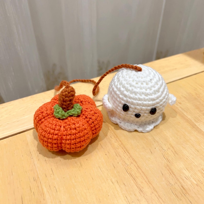 May include: Crocheted Halloween decorations: an orange pumpkin with a brown stem and green leaves, and a white ghost with black eyes and a small mouth. Both have brown yarn loops.