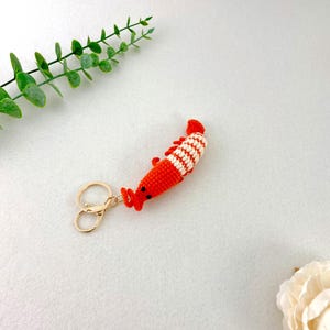 May include: A handmade crochet shrimp keychain in bright orange and white stripes. The shrimp has small black eyes and a gold-colored clasp. The keychain is a fun accessory.