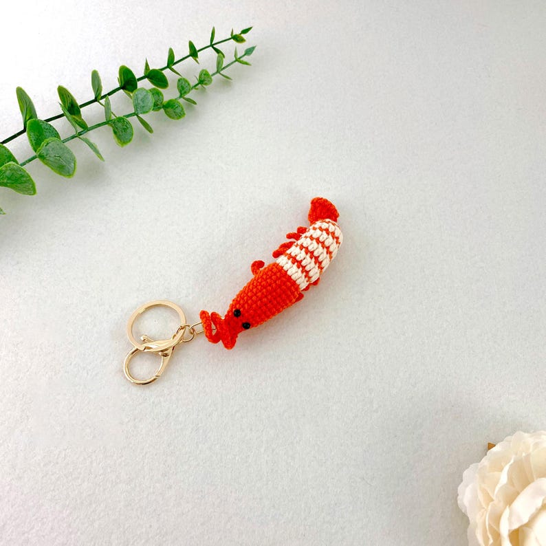 May include: A handmade crochet shrimp keychain in bright orange and white stripes. The shrimp has small black eyes and a gold-colored clasp. The keychain is a fun accessory.