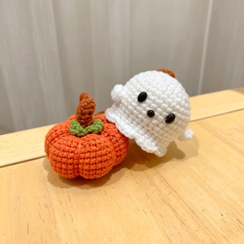May include: A handmade crochet Halloween decoration featuring a small orange pumpkin with a brown stem and green leaves, next to a white ghost with black button eyes. The ghost has a small brown patch on top.