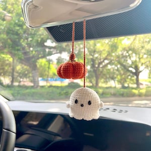 May include: A handmade car charm featuring a crocheted pumpkin and ghost. The pumpkin is orange, and the ghost is white with black eyes. The charm hangs from a brown string, adding a touch of Halloween decor to the car's rearview mirror.