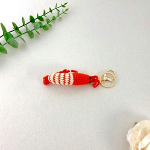 May include: A handmade crochet shrimp keychain. The shrimp is primarily orange with white stripes and red accents. It has a gold-colored metal clasp for attaching to keys or bags. The keychain is a whimsical accessory.