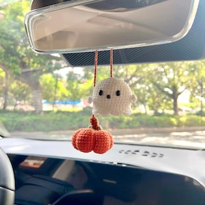 May include: A handmade Halloween car charm featuring a white crochet ghost and an orange crochet pumpkin. The ghost has black button eyes and a small mouth. The pumpkin has a green stem. The charm hangs from a rearview mirror.