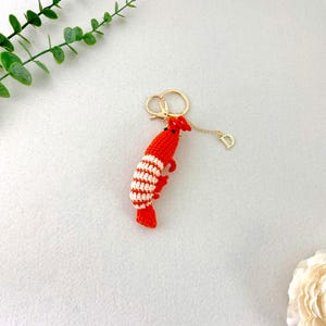 May include: A handmade red and white crochet shrimp keychain with a gold-colored keyring and a small charm. The shrimp has a detailed design with red and white stripes. The keychain is on a light gray surface.