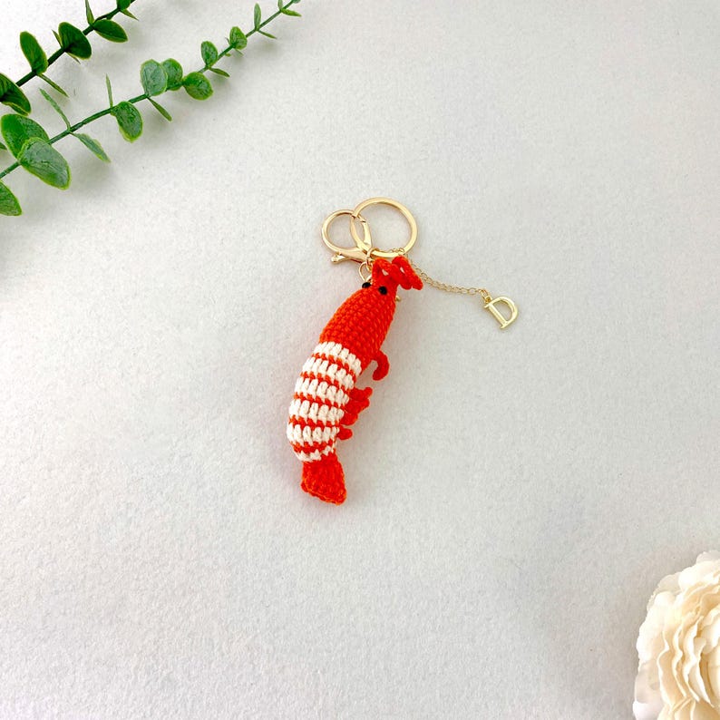May include: A handmade red and white crochet shrimp keychain with a gold-colored keyring and a small charm. The shrimp has a detailed design with red and white stripes. The keychain is on a light gray surface.