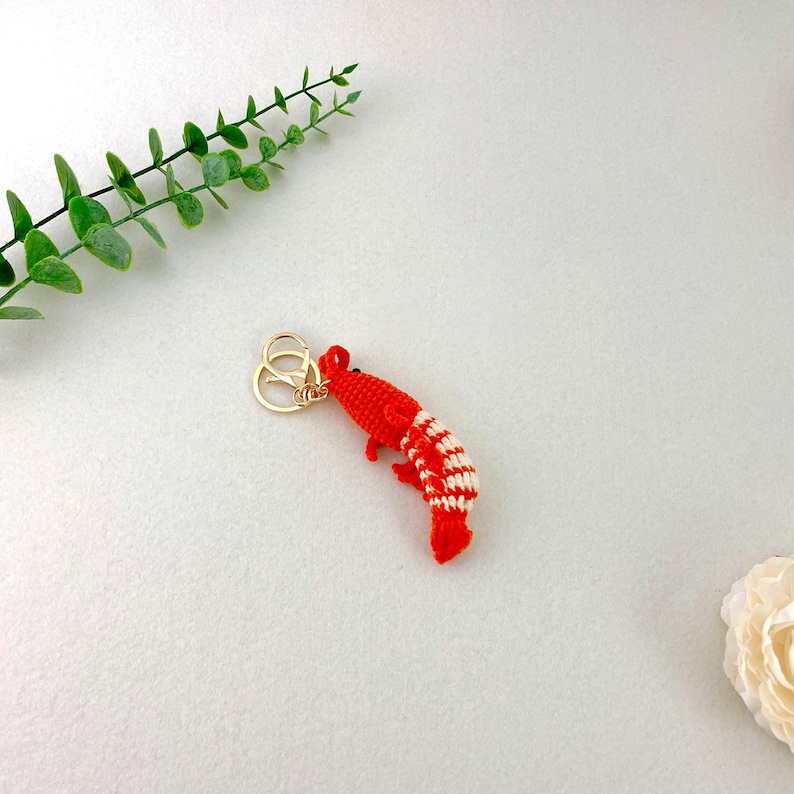 May include: A handmade red and white shrimp keychain with a gold-colored clasp. The shrimp is crocheted with a detailed pattern, and the keychain is set against a white background with green foliage.