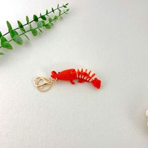 May include: A handmade crochet shrimp keychain. The shrimp is primarily bright red with white and red striped detailing on the tail. It has a gold-colored metal clasp for attaching to keys or bags. The keychain is a unique, handcrafted accessory.