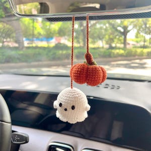 May include: Crocheted Halloween car charm featuring a small orange pumpkin and a white ghost with black eyes. The pumpkin has a green stem. The charm hangs from a brown string, perfect for seasonal decor.