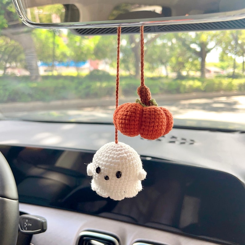May include: Crocheted Halloween car charm featuring a small orange pumpkin and a white ghost with black eyes. The pumpkin has a green stem. The charm hangs from a brown string, perfect for seasonal decor.