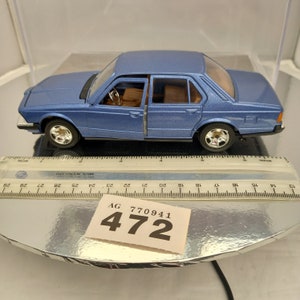 May include: A blue die-cast model car of a BMW 7-series saloon. The car is on a silver surface with a white tag that reads "AG 770941 472".