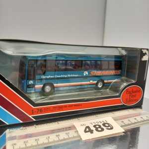 Puede incluir: Un modelo de autobús de fundición a presión a escala 1/76, de color azul y naranja, con las palabras "Carefree Coaching Holidays..." y "Shearings" en el lateral. El autobús está en una caja de plástico transparente con un fondo de rayas rojas y negras. La caja también tiene las palabras "Exclusive First Editions" en ella.