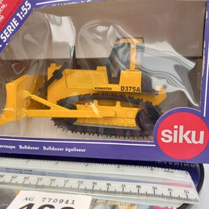 May include: A yellow Komatsu D375A bulldozer model in a clear plastic case. The model has black tracks and a blade. The box has the text "SERIE 1:55" and the brand "siku".